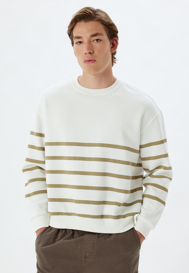 Sudadera blanca con rayas horizontales en color verde oliva, con cuello redondo y puños acanalados. Combinada con pantalones marrones.