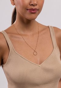 Goldfarbene Halskette mit einem runden Anhänger; getragen über einem beigefarbenen, gerippten Tanktop. Der Stoff hat ein strukturiertes, vertikales Muster.