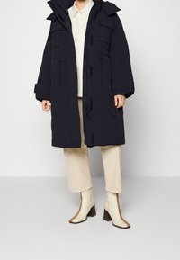 Giacca a vento nera con grandi tasche e cappuccio con cordino. Materiale spesso con finitura testurizzata. Abbinata a pantaloni color crema e stivali con tacco.