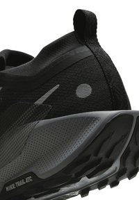 Chaussure de trail Nike noire et grise avec semelle texturée, boucle de traction au talon et détail circulaire réfléchissant sur le côté.