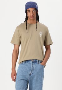 T-shirt khaki à manches courtes avec un logo rond sur la poitrine, associé à un jean bleu clair ample. Le modèle porte un bonnet rayé bleu.