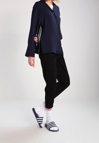 Camisa de botones azul marino con aberturas laterales, combinada con pantalones negros ajustados. Lleva calcetines blancos con sandalias deslizantes de rayas azules. Un atuendo simple y casual.