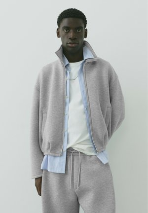 Jeune homme portant une veste zippée grise, un pantalon de jogging assorti, une chemise bleue claire à boutons et un t-shirt blanc sur un fond clair uni.