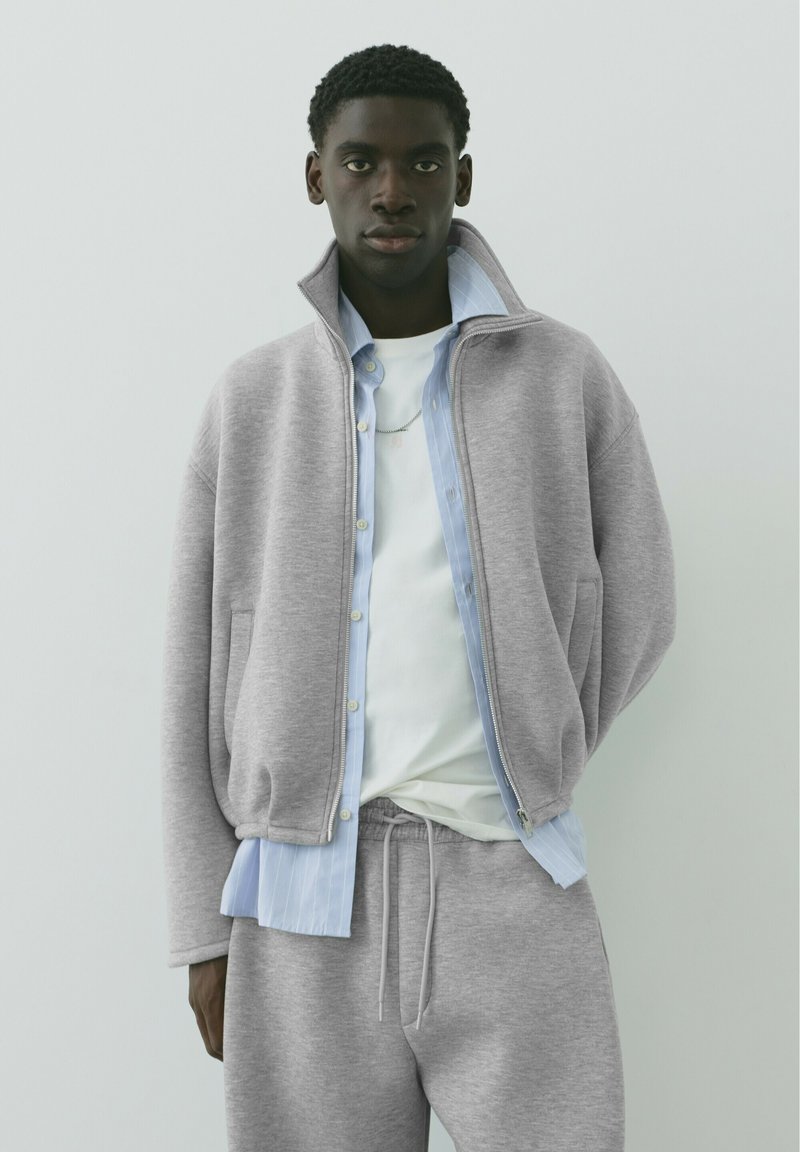 Jeune homme portant une veste zippée grise, un pantalon de jogging assorti, une chemise bleue claire à boutons et un t-shirt blanc sur un fond clair uni.