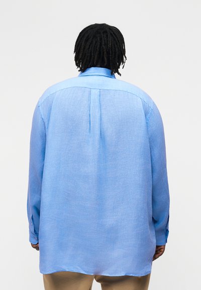 Asmuo su tamsiais pintais plaukais, vilkintis laisvai krentančią šviesiai mėlynos spalvos ilgomis rankovėmis marškinių ir smėlio spalvos kelnių porą, matomas iš už nugaros priešais vientisą foną.