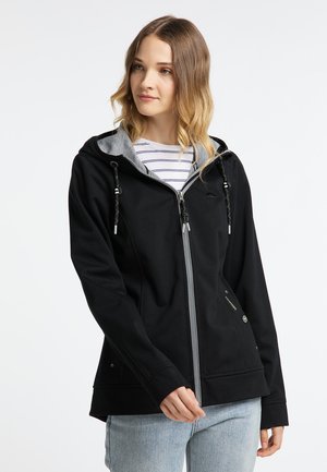 Junge Frau trägt einen schwarzen Kapuzenpullover mit Reißverschluss über einem gestreiften Shirt und hellblauen Jeans und steht vor einem weißen, schlichten Hintergrund.