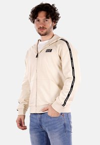 Felpa con zip color beige realizzata in tessuto morbido, con un patch logo nero e strisce laterali a contrasto. Tasche anteriori e cappuccio con cordino.