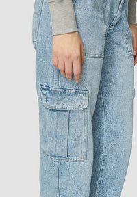 Lichtblauwe denim cargo joggers met zijzakken en zichtbare stiksels. De stof heeft een licht gestructureerde afwerking. Grijze shirt met lange mouwen is zichtbaar.