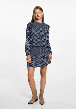 Marineblaues Kleid mit weißem geometrischem Muster, geraffter Textur, langen Ärmeln und Rüschendetails an den Schultern. Tragen mit hellbeigen Knöchelstiefeln.