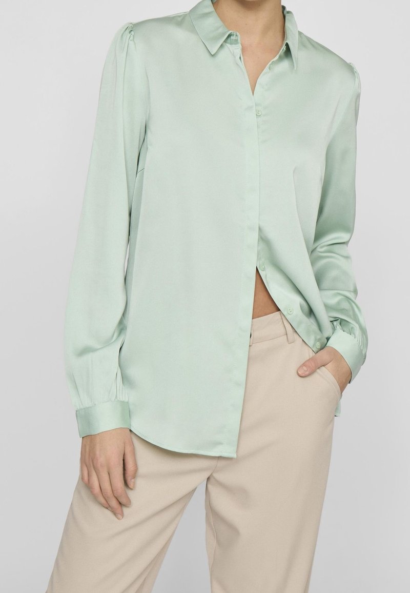 Femme portant un chemisier satiné vert clair à manches longues avec boutons et un pantalon beige, une main dans la poche, sur un fond neutre.