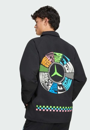 Person iført sort jakke med flerfarvet cirkulært grafik og skakternet mønster på ryggen, stående mod en ensfarvet lys baggrund.