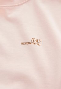 Camiseta de algodón de color rosa claro con texto impreso que dice "ITALIA Mar Mediterráneo" en marrón. Textura suave y diseño de cuello redondo.
