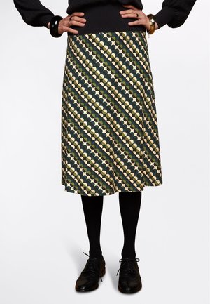 Kobieta ubrana w spódnicę do kolan z ukośnym wzorem zielonych, beżowych i czarnych okręgów, czarne rajstopy, czarne buty i czarny sweter.