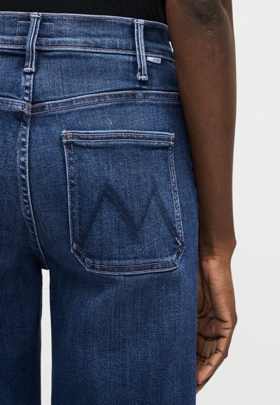 Κοντινό πλάνο ατόμου που φοράει μπλε τζιν με κεντητό σχεδιασμό "M" στην πίσω τσέπη, στέκεται μπροστά σε απλό φόντο.