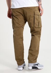 Pantaloni cargo in tessuto verde oliva, con multiple tasche laterali, design a gamba dritta e dettagli di cucitura. Indossati con sneaker grigie.