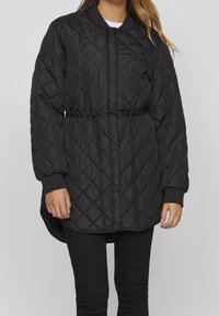 Svart quiltad puffjacka med dragsko i midjan, långa ärmar och ribbstickade manschetter; har en rundad halsringning och en något förlängd bakdel.