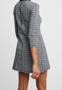 Robe à carreaux noir et blanc avec des manches longues, une taille cintrée et une jupe évasée. Dispose d'une fermeture éclair au dos et d'une texture lisse.
