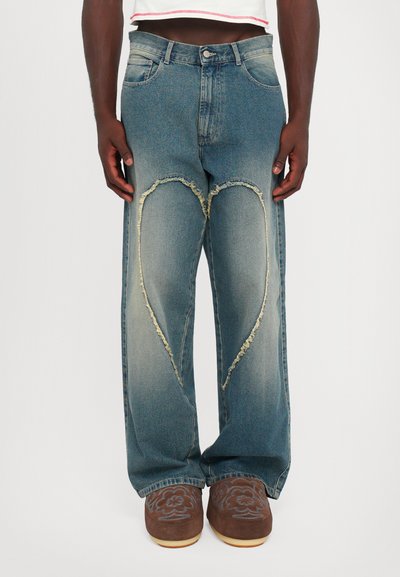 Persoon in blauwe jeans met grote hartvormige rafelige patchs op de voorkant en bruine geborduurde instappers.
