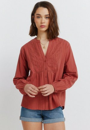 Ung kvinde iført en rustrød broderet bluse og opsmøgede denimshorts, stående mod en ensartet lys baggrund.