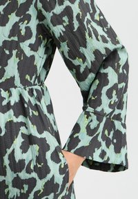 Material de culoare albastru deschis și negru, cu un model texturat și ondulat. Prezintă un design cu mânecă clopot și o talie ajustată, având o buzunar lateral.