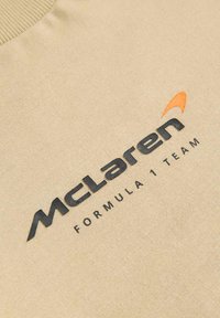 Béžová mikina s žebrovaným výstřihem, na které je černý text "McLaren Formula 1 Team" a oranžová akcentní logo. Měkká textura, ležérní design.