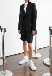 Svart dubbelknäppt kavaj över en vit skjorta, kombinerad med svarta shorts och vita sneakers. Slät tyg, skräddarsydd passform och minimalistisk design.