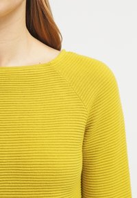 Haut en tricot côtelé jaune avec un col bateau et des manches raglan, présentant une surface texturée et une coupe près du corps.