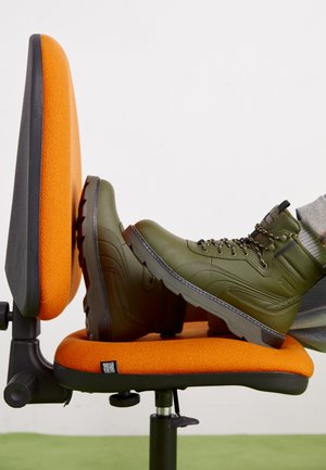 Bottes vert olive avec accents noirs et œillets en métal, reposant sur une chaise rembourrée orange, mettant en valeur une semelle texturée et un col rembourré.