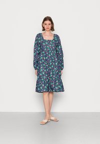 Vestido floral em azul marinho com padrões verdes e cor-de-rosa. Apresenta um decote quadrado, mangas compridas e uma saia em camadas. Acompanhado por sapatos bejes planos.