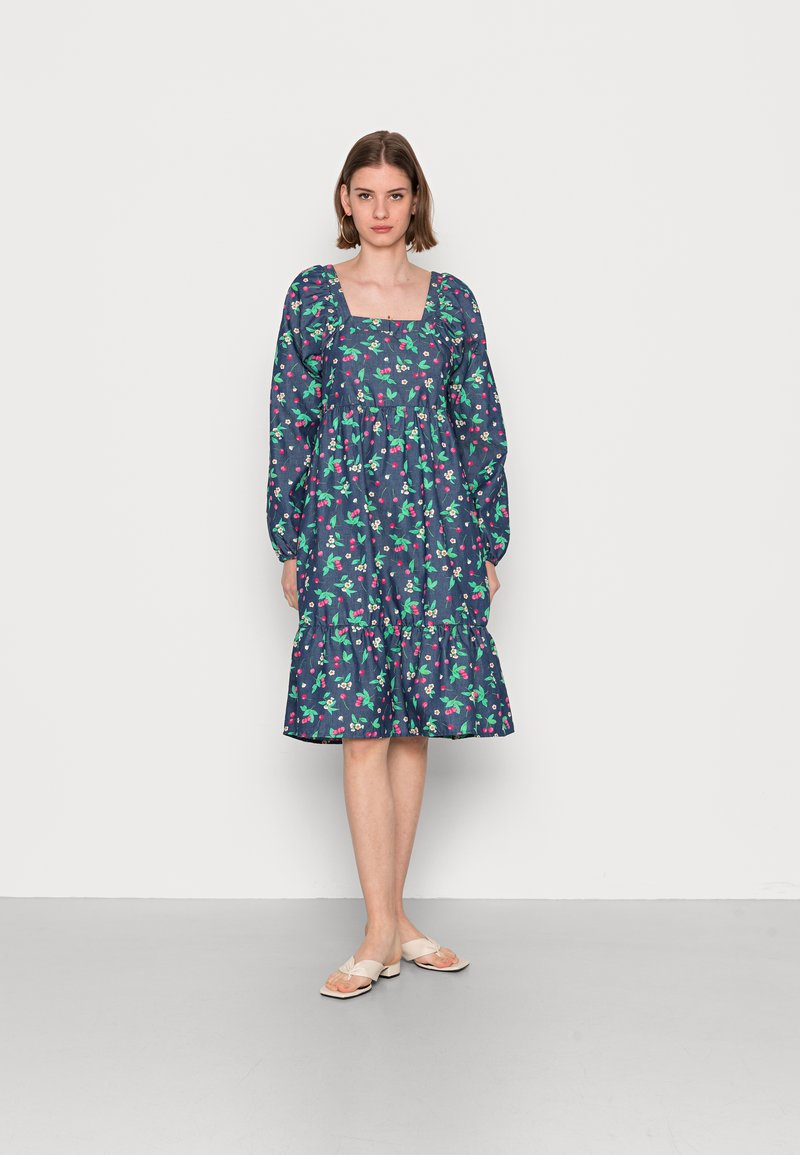 Vestido floral em azul marinho com padrões verdes e cor-de-rosa. Apresenta um decote quadrado, mangas compridas e uma saia em camadas. Acompanhado por sapatos bejes planos.