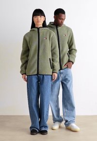 Twee modellen staan naast elkaar, gekleed in groene fleece hoodies met rits, brede blauwe jeans en casual schoenen, tegen een effen witte achtergrond.