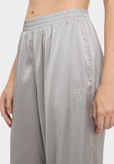 Grijze satijnen broek met een elastische tailleband en zijplooien. Heeft een witte "7" logo op het onderbeen. Soepele textuur.