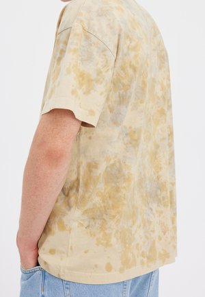 Osoba nosząca beżową i brązową koszulkę z krótkim rękawem w stylu tie-dye oraz jasnoniebieskie jeansy, stojąca z lewą ręką w kieszeni, zwrócona bokiem.