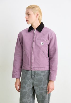 Giacca di cotone viola con colletto in velluto a coste neri, chiusura frontale con zip e taschino al petto. Indossata con pantaloni grigi a fantasia, caratterizzati da una vestibilità comoda.