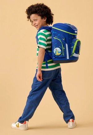 Smiling child with curly hair walking, wearing green striped shirt, blue jeans, white sneakers, and a large blue backpack with lime green accents.