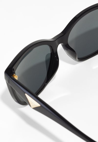Black sunglasses with dark lenses, angular frames, and gold accent on the temples. Smooth plastic material, modern design with bold shape.