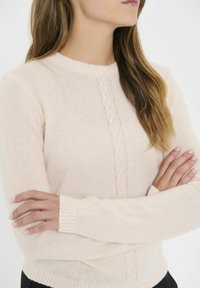 Jasnoróżowy sweter wykonany na drutach z okrągłym dekoltem, prążkowanymi mankietami i subtelnym wzorem w warkocze na przodzie. Miękka tekstura, dopasowany krój.