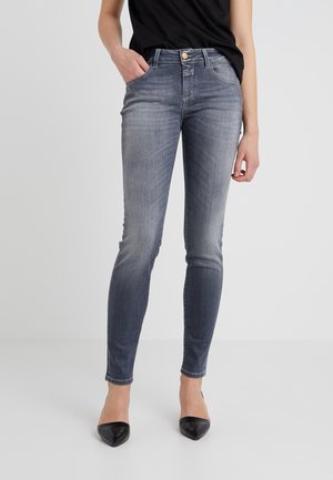 Kobieta ubrana w dopasowane, szare, sprane dżinsy, czarne szpilki z ostrym noskiem i czarną bluzkę, stojąca na jasnym, jednolitym tle.