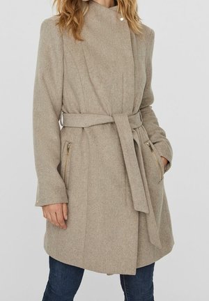 Žena v béžovém kabátu ke kolenům se zavazováním v pase, se zipovými bočními kapsami a dlouhými rukávy, v kombinaci s tmavě modrými džínami.