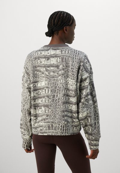 Kvinne med flettet hår iført en oversized langermet topp med grå og hvit krokodilleskinnmønster, kombinert med mørke leggings, snudd bort.