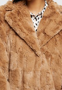 Fuskpälsjacka i ljust brunt med en texturerad, tovlig yta och en bred krage. Har en lös passform och lager-på-lager tygdesign.