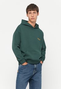 Felpa verde realizzata in tessuto texturizzato, con una tasca a marsupio e logo arancione sul petto. Abbinata a jeans di denim blu.