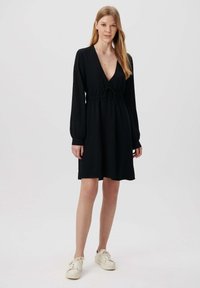 Schwarzes Kleid mit langen Ärmeln, tiefem V-Ausschnitt, taillierter Taille, aus einem strukturierten Stoff; kombiniert mit cremefarbenen Sneakers.