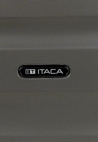 Logotipo ITACA en negro y plata sobre una superficie de tela gris oscuro texturizada, probablemente el exterior de un equipaje o bolso.