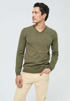 Pull en maille légère de couleur vert olive avec un col en V, des poignets et un ourlet côtelés. Associé à un pantalon beige, sans motifs ni logos visibles.