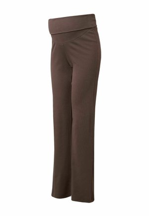 Donna incinta che indossa una maglia premaman aderente marrone scuro e pantaloni a zampa, in piedi davanti a uno sfondo beige uniforme.