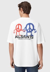 Hombre vestido con una camiseta blanca con signos de paz rojos y azules, sosteniendo las manos por encima del texto "MENSAJE DE ALLSAINTS A UN FUTURO" en la parte de atrás.