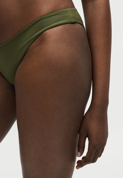 Κοντινό πλάνο στο πάνω μέρος του μηρού και το χέρι ενός ατόμου που φοράει εσώρουχα μπικίνι σε ελιά πράσινο χρώμα κατάλληλα τοποθετημένα σε ένα απλό ανοιχτόχρωμο φόντο.