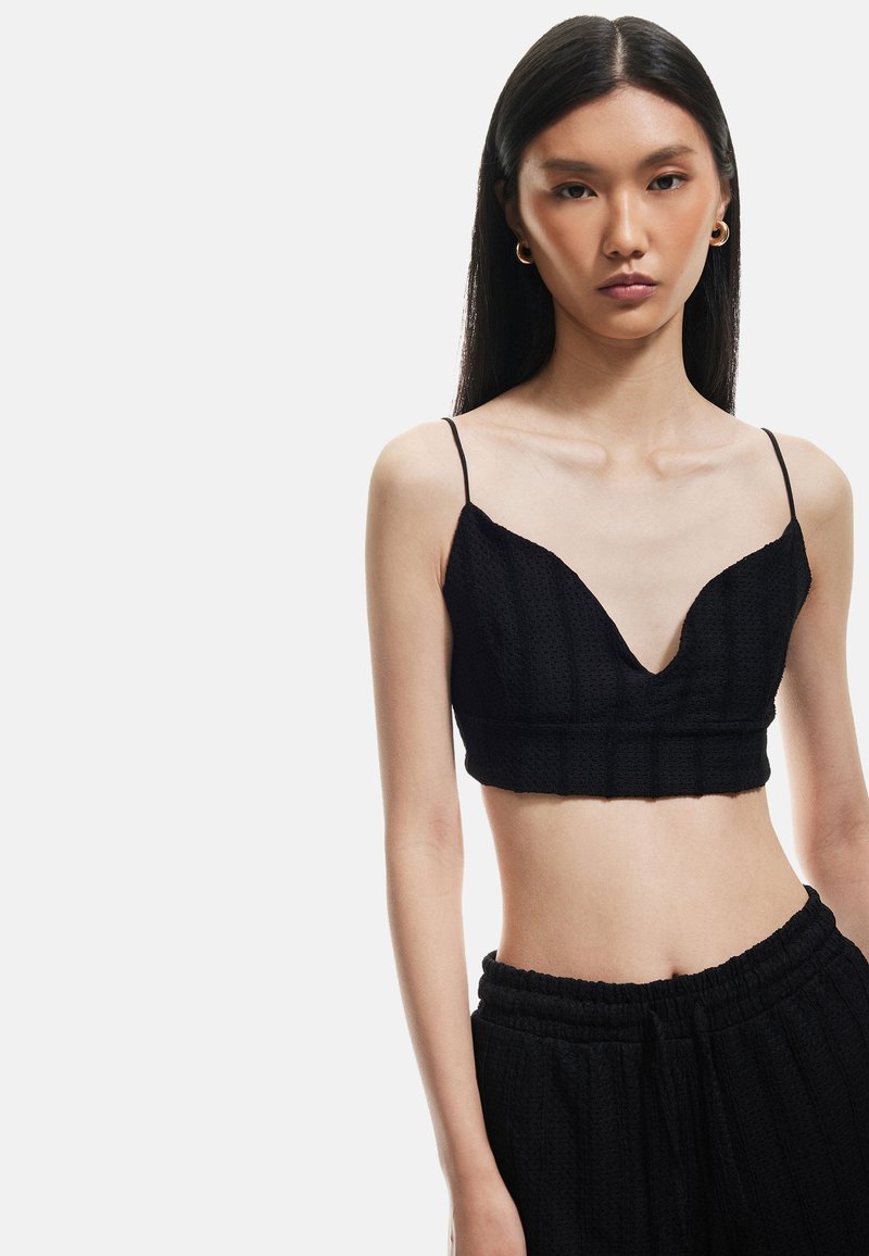 Sort, cropped bralette med en dyb V-udskæring, tekstureret stof og tynde spaghettistropper. Sættes sammen med matchende sorte bukser med elastisk talje.