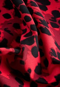 Tissu rouge à texture douce avec des taches de léopard noires irrégulières, présenté avec des plis créant de la profondeur et des ombres.